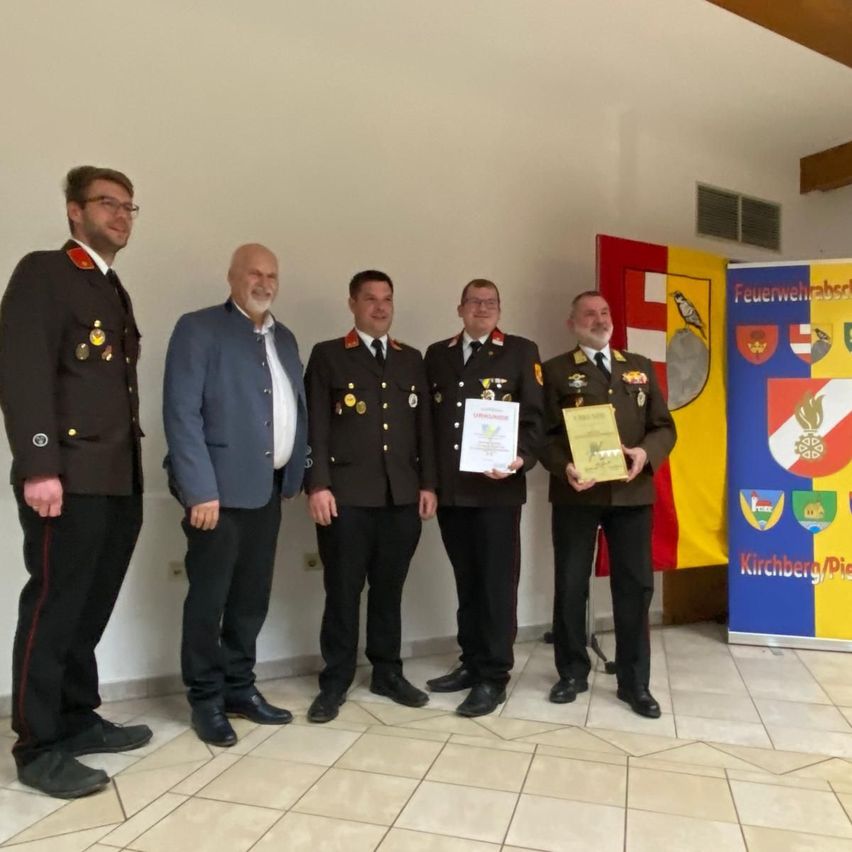 Fünf Männer in Uniformen stehen zusammen, zwei halten Urkunden. Sie lächeln für ein Foto vor einer weißen Wand mit einem bunten Banner.