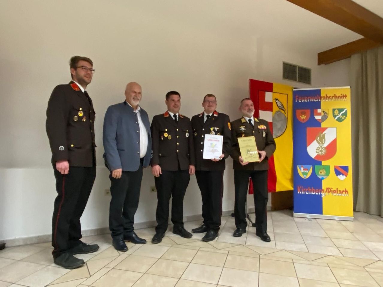 Fünf Männer in Uniformen stehen zusammen, zwei halten Urkunden. Sie lächeln für ein Foto vor einer weißen Wand mit einem bunten Banner.