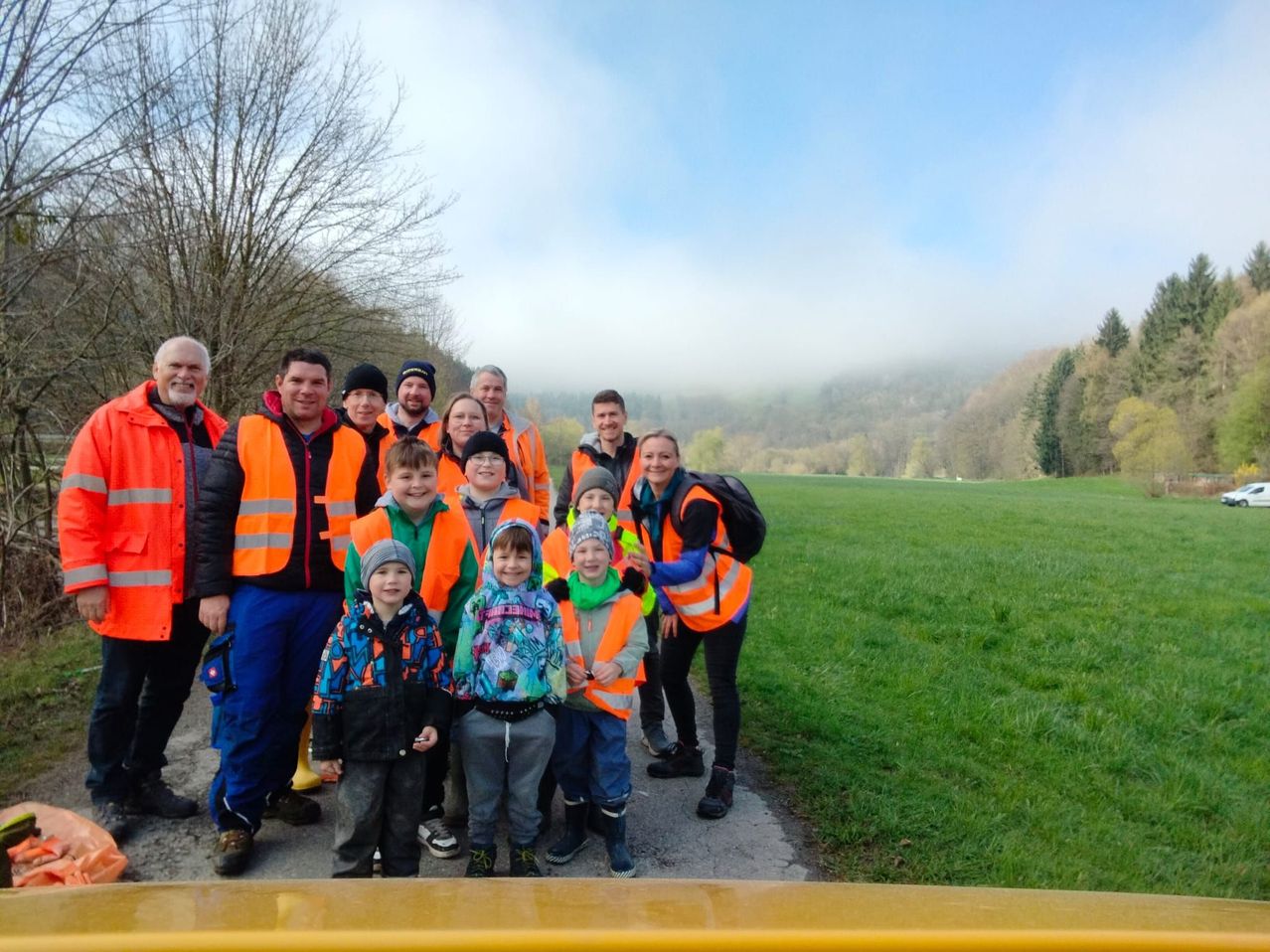 Eine Gruppe von Erwachsenen und Kindern in leuchtend orangefarbenen Westen steht in einem Feld mit einem nebligen Hintergrund.