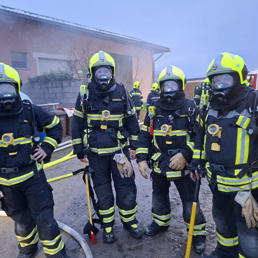 Eine Gruppe von Feuerwehrleuten in voller Ausrüstung steht vor einem Gebäude. Sie sind mit Helmen, Masken und Handschuhen ausgestattet. Hinter ihnen steht ein Feuerwehrwagen.
