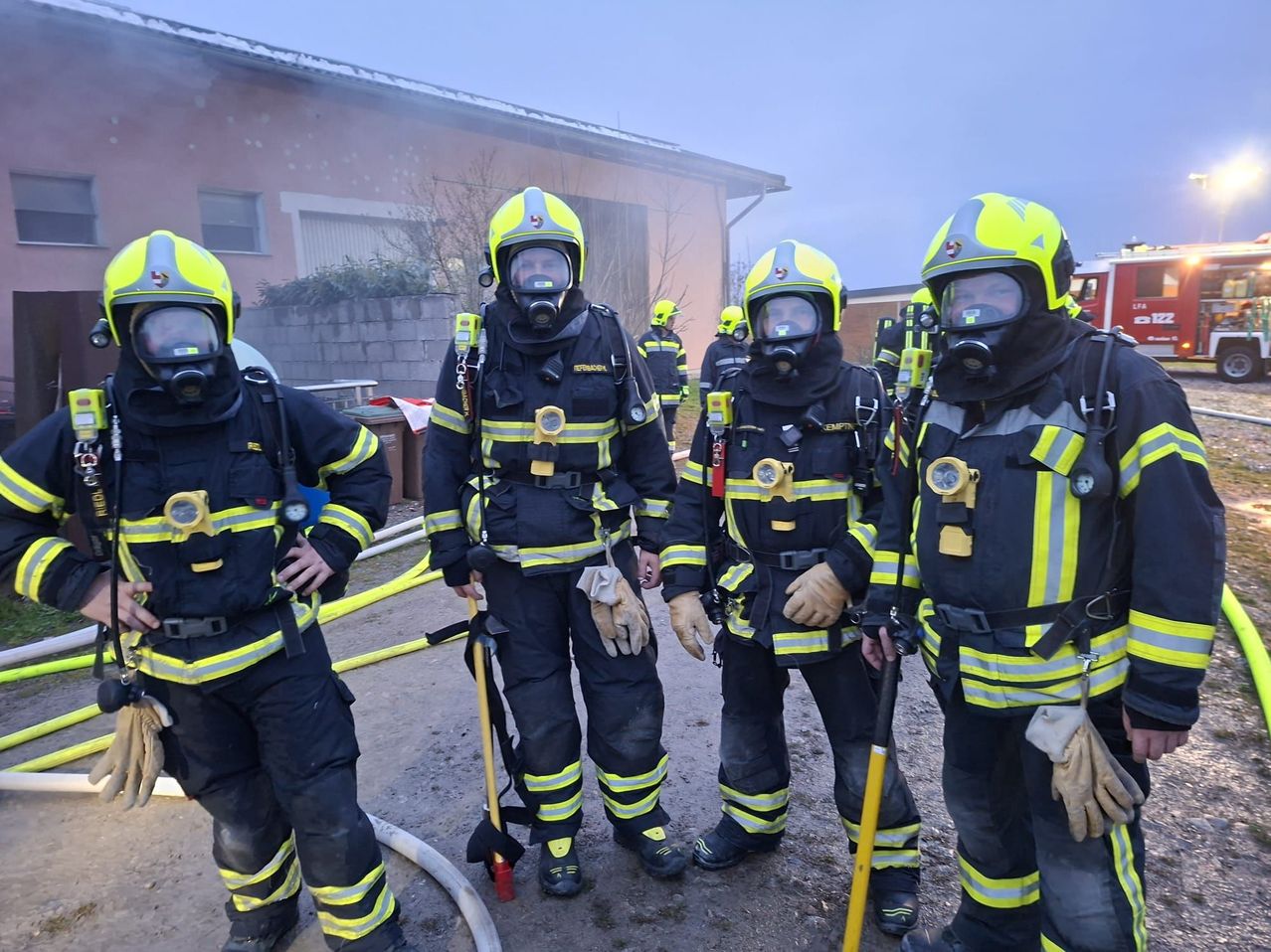 Eine Gruppe von Feuerwehrleuten in voller Ausrüstung steht vor einem Gebäude. Sie sind mit Helmen, Masken und Handschuhen ausgestattet. Hinter ihnen steht ein Feuerwehrwagen.