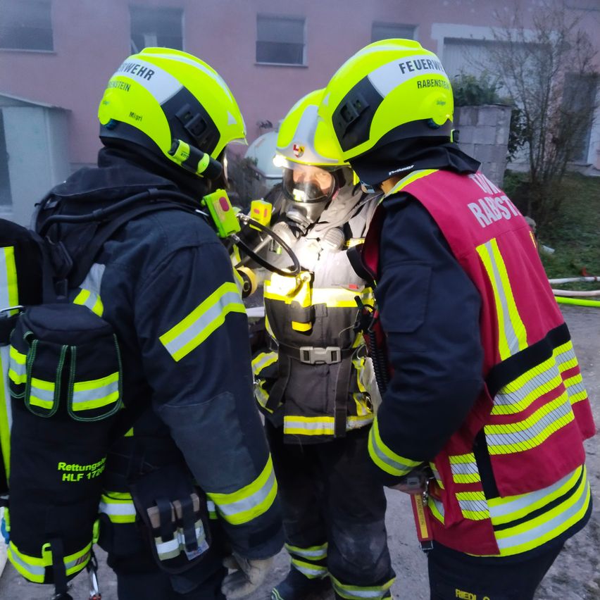 Drei Feuerwehrleute stehen vor einem Gebäude, einer von ihnen trägt eine Gasmaske. Sie tragen gelbe und schwarze Helme.