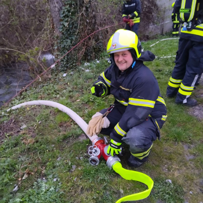 Ein Feuerwehrmann kniet auf dem Gras, hält einen Feuerwehrschlauch und lächelt, mit einem Bach im Hintergrund.
