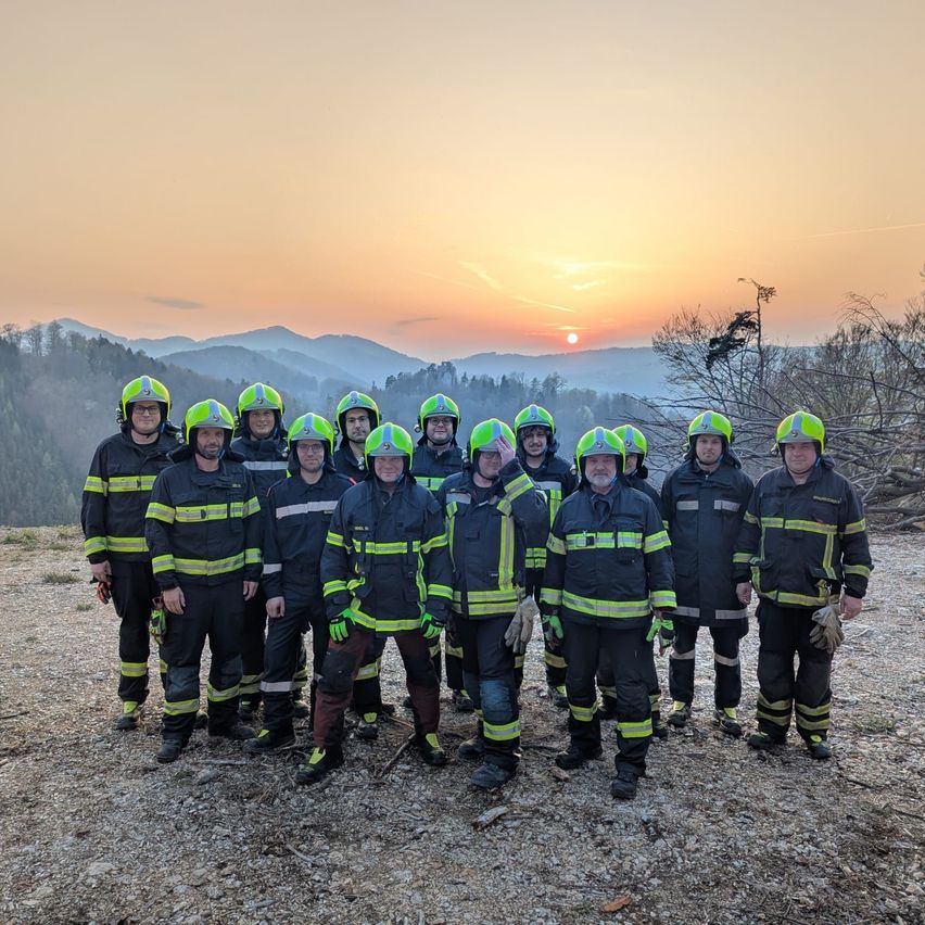 Eine Gruppe von Feuerwehrleuten in Schutzausrüstung posiert für ein Foto auf einem Berg bei Sonnenuntergang mit Blick auf die Berge in der Ferne.