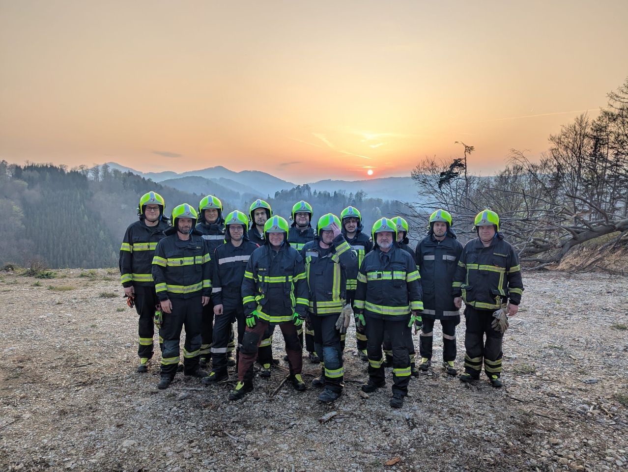 Eine Gruppe von Feuerwehrleuten in Schutzausrüstung posiert für ein Foto auf einem Berg bei Sonnenuntergang mit Blick auf die Berge in der Ferne.
