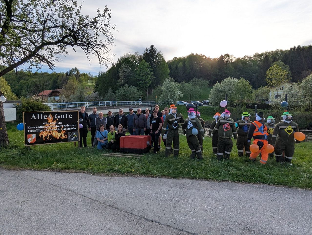 Eine Gruppe von Menschen in Kostümen und Masken posiert für ein Foto neben einem Schild, mit einer Brücke und Bäumen im Hintergrund.