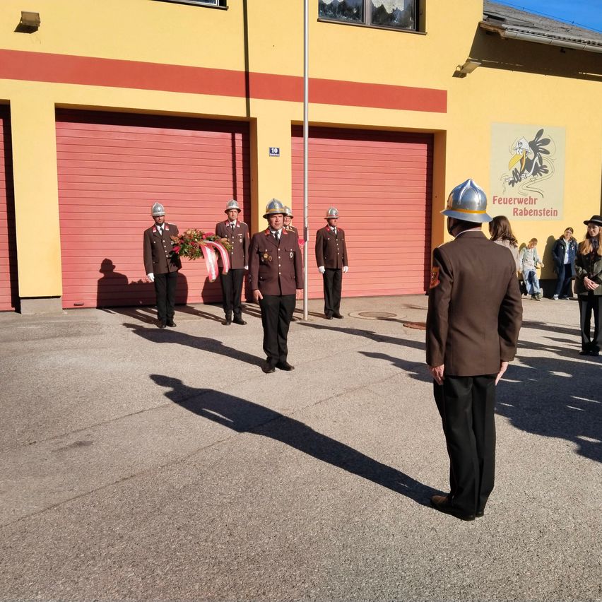 Fünf Feuerwehrleute in Uniform stehen vor einer Feuerwache, mit einem Kranz auf dem Boden und Zuschauern dahinter.