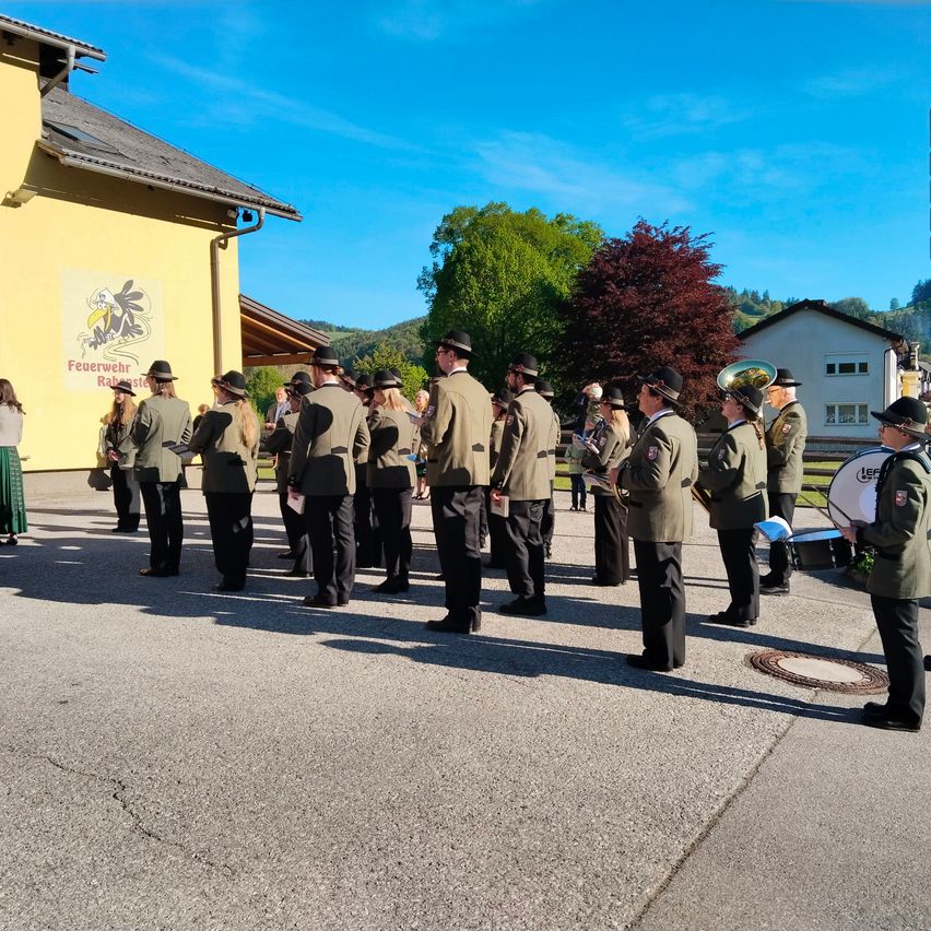 Eine Blaskapelle in grünen Uniformen steht in einer Reihe und hält Instrumente. Sie stehen vor einem Gebäude mit einem Schild, auf dem 'Feuerwehr' steht. Eine Frau in einem grünen Kleid steht links.