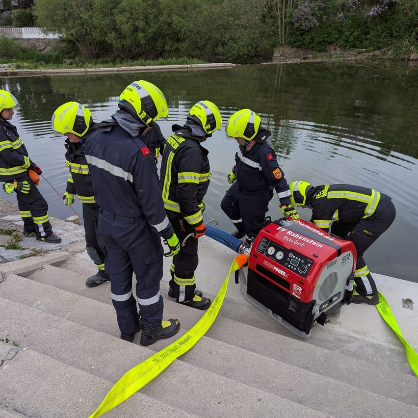 Feuerwehrleute in gelben Helmen bereiten einen Generator zur Verwendung in der Nähe eines Gewässers vor. Die Gruppe steht auf Stufen, einer beugt sich über den Generator.