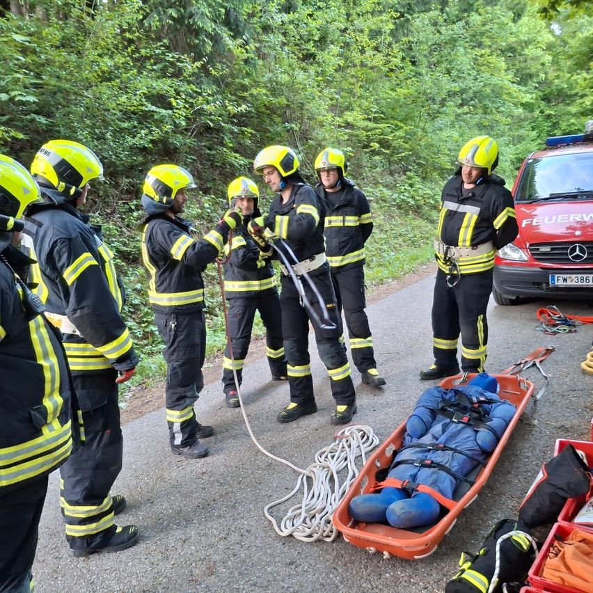 Feuerwehrleute in gelben Helmen üben eine Seilrettung auf einer Straße. Ein Dummy liegt in einer orangefarbenen Trage. Ein roter Mercedes-Van ist geparkt.