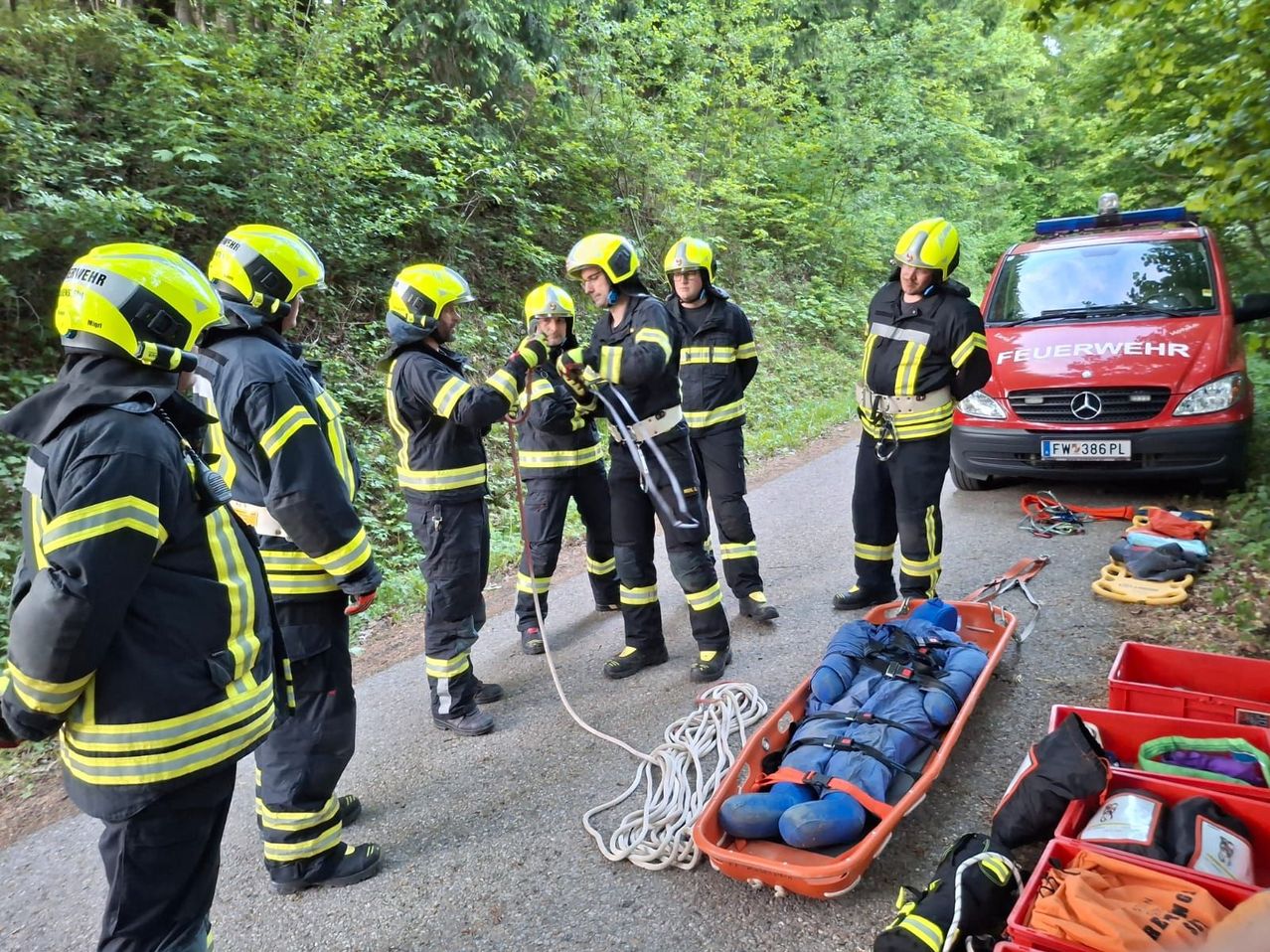 Feuerwehrleute in gelben Helmen üben eine Seilrettung auf einer Straße. Ein Dummy liegt in einer orangefarbenen Trage. Ein roter Mercedes-Van ist geparkt.