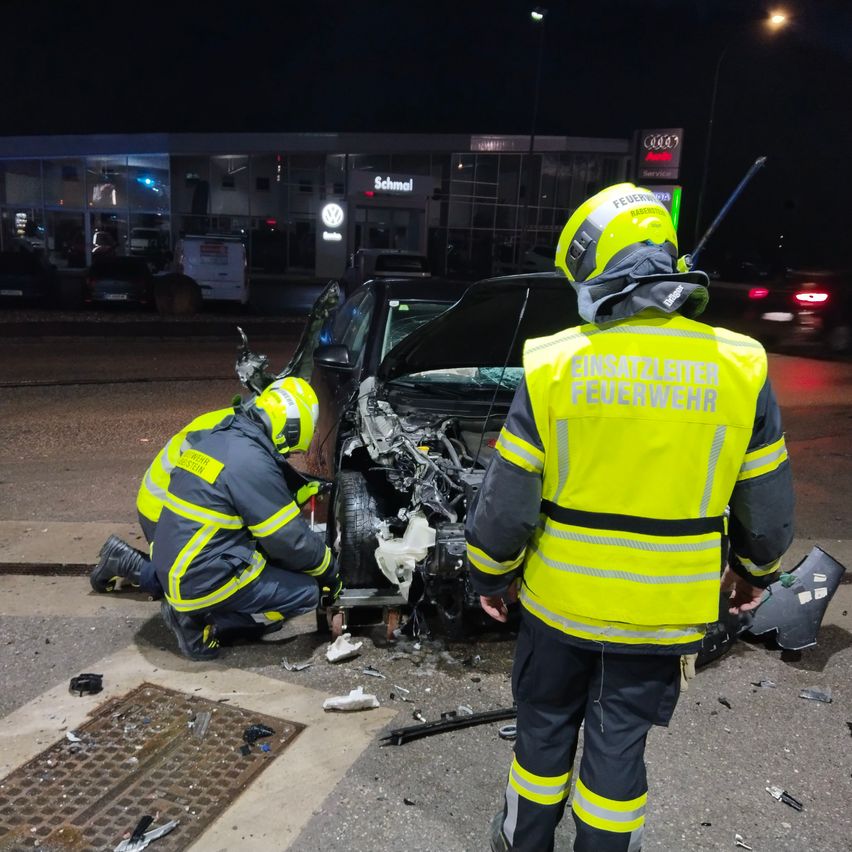 Zwei Feuerwehrleute in reflektierenden Westen inspizieren ein beschädigtes Auto am Straßenrand bei Nacht, mit einer Tankstelle im Hintergrund.
