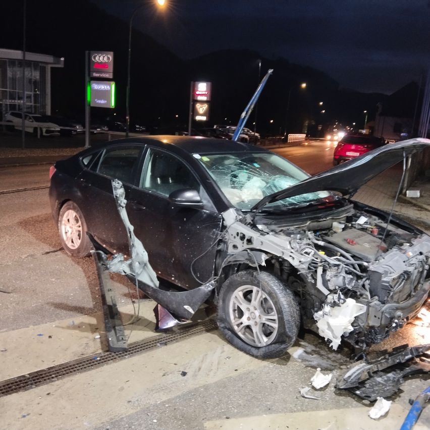 Ein beschädigtes schwarzes Auto steht nachts am Straßenrand, die Motorhaube ist geöffnet und die Vorderseite ist stark beschädigt. Das Auto befindet sich neben einem Bürgersteig. Dahinter ist ein Audi-Händler zu sehen, und im Hintergrund sind Straßenlaternen und andere Fahrzeuge zu erkennen.