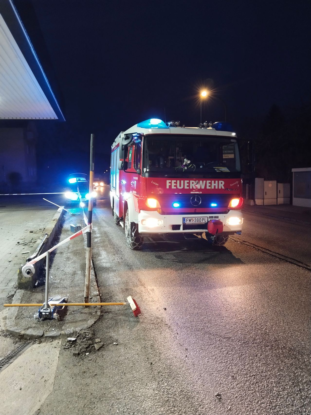 Ein Feuerwehrauto steht nachts mit eingeschalteten Lichtern am Straßenrand. In der Nähe befindet sich ein Auto mit blauen Lichtern und ein Pfosten mit einem weiß-roten Band.