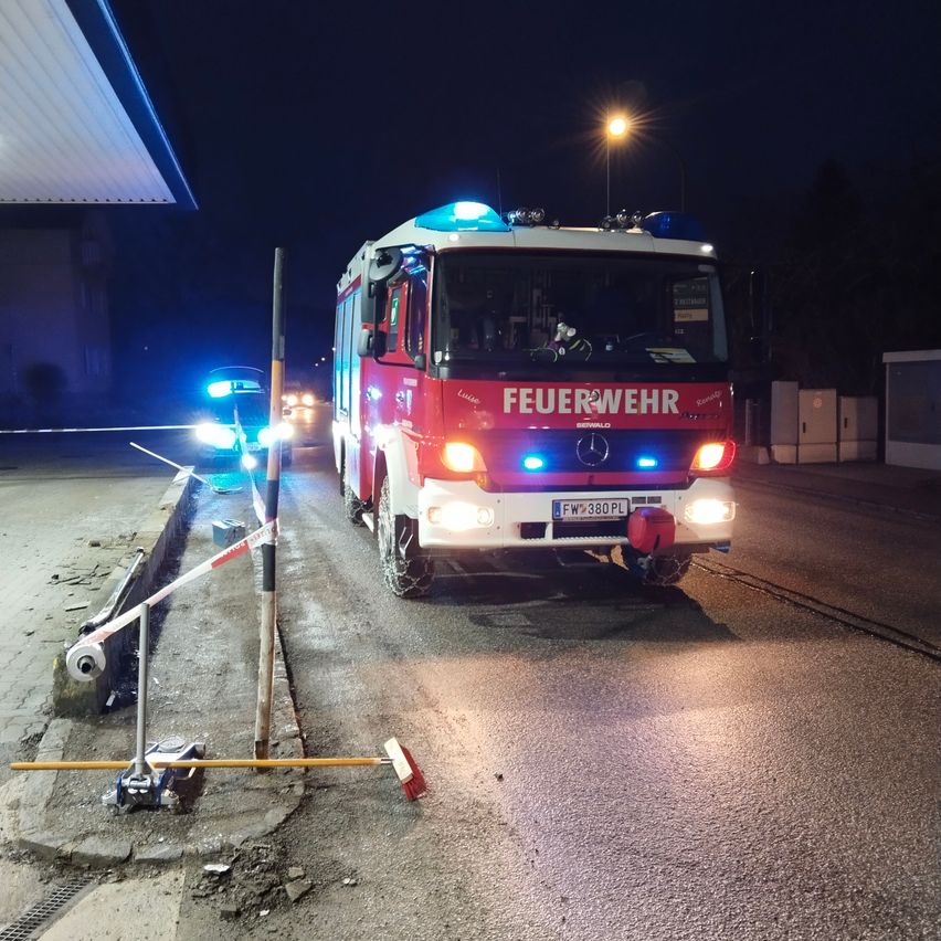 Ein Feuerwehrauto steht nachts mit eingeschalteten Lichtern am Straßenrand. In der Nähe befindet sich ein Auto mit blauen Lichtern und ein Pfosten mit einem weiß-roten Band.