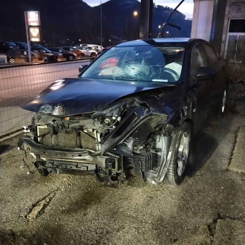 Ein schwarzer Wagen mit schweren Frontschäden steht am Straßenrand. Die Windschutzscheibe ist zersplittert und der Stoßfänger ist abgerissen. Hinter einer Barriere sind weitere Autos geparkt.