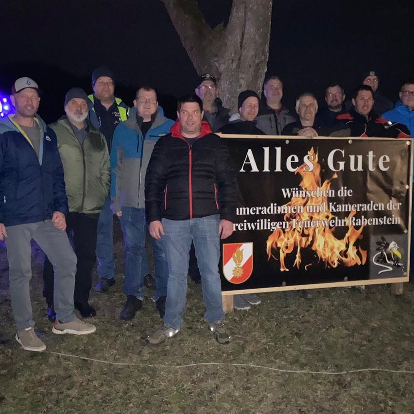 Eine Gruppe von Männern steht vor einem Banner mit einem Feuerbild, ein Mann hält es, andere dahinter, tragen Mützen und Jacken, möglicherweise bei einer Nachtveranstaltung.