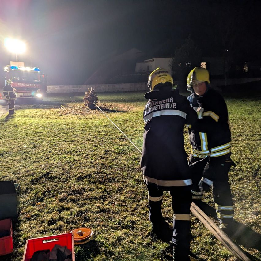 Zwei Feuerwehrleute in gelben Helmen stehen nachts auf einem Feld und halten eine Feuerwehrschläuche. Ein Feuerwehrauto ist in der Nähe geparkt, mit eingeschalteten Lichtern. Ein Gebäude ist im Hintergrund sichtbar.