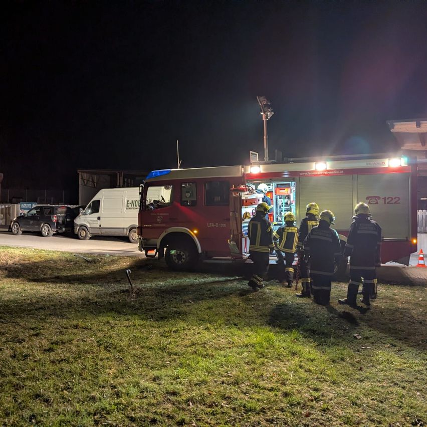 Feuerwehrleute stehen um einen roten Feuerwehrwagen mit eingeschalteten Lichtern herum, der nachts auf einem Rasenparkplatz geparkt ist.