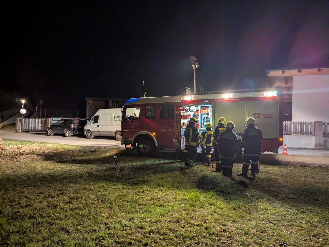 Feuerwehrleute stehen um einen roten Feuerwehrwagen mit eingeschalteten Lichtern herum, der nachts auf einem Rasenparkplatz geparkt ist.