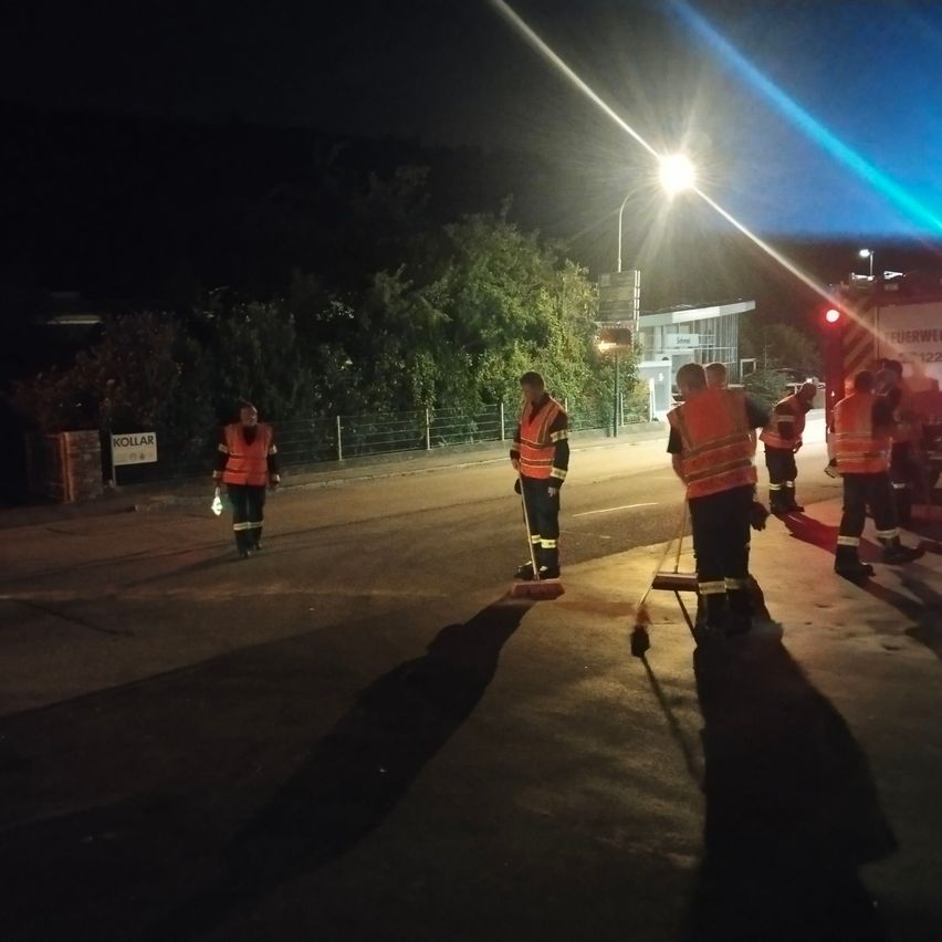 Eine Gruppe von Feuerwehrleuten in Warnwesten reinigt die Straße bei Nacht. Sie werden von einer Straßenlaterne und einem Feuerwehrwagen beleuchtet.