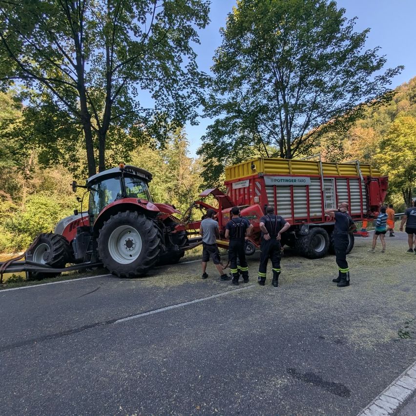 Ein roter Traktor ist am Straßenrand stecken geblieben, mit einem roten Anhänger. Mehrere Personen in Sicherheitsausrüstung stehen darum herum, möglicherweise um zu helfen.