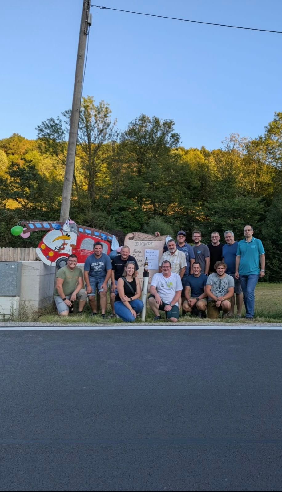 Eine Gruppe von Menschen posiert für ein Foto vor einer bemalten Wand und einem dekorierten Auto an einem sonnigen Tag.