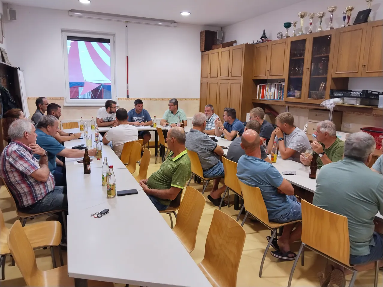 Eine Gruppe von Männern in Freizeitkleidung sitzt um einen Tisch in einem Raum herum, mit Flaschen und Gläsern auf dem Tisch. Sie scheinen an einer Besprechung oder Diskussion teilzunehmen.