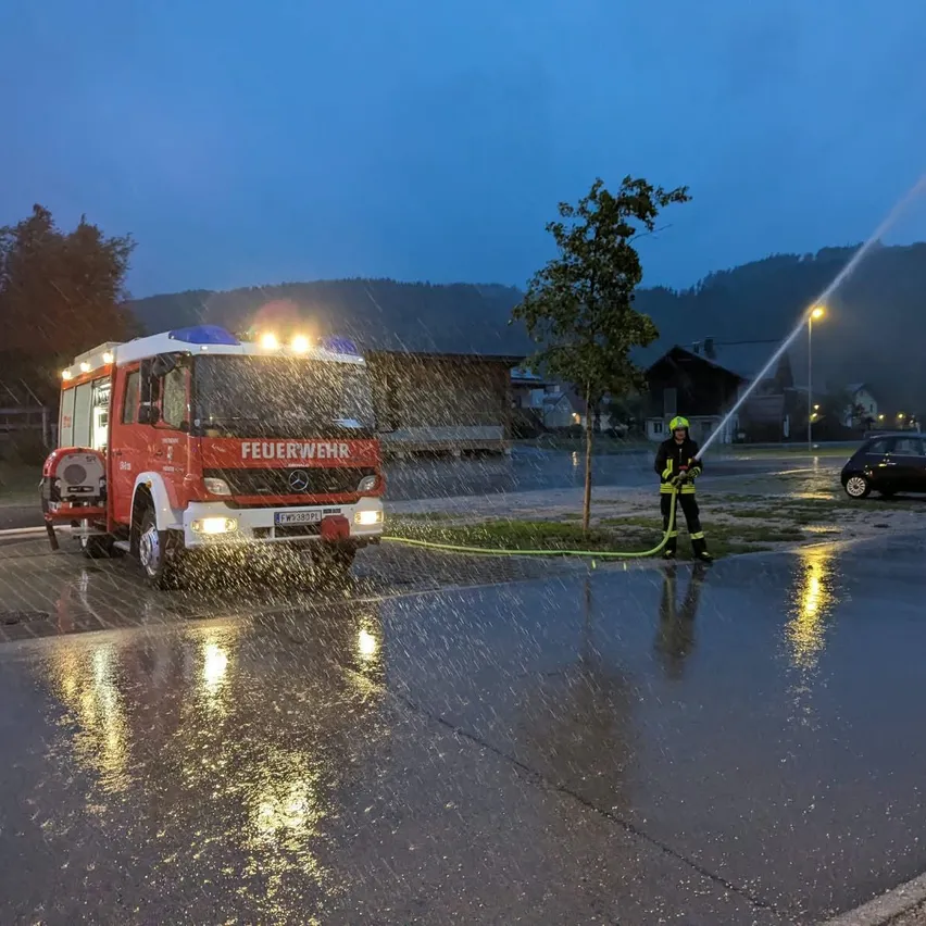 Ein Feuerwehrmann in einem gelben Helm sprüht Wasser aus einem Feuerwehrwagen bei Regen in der Nacht.
