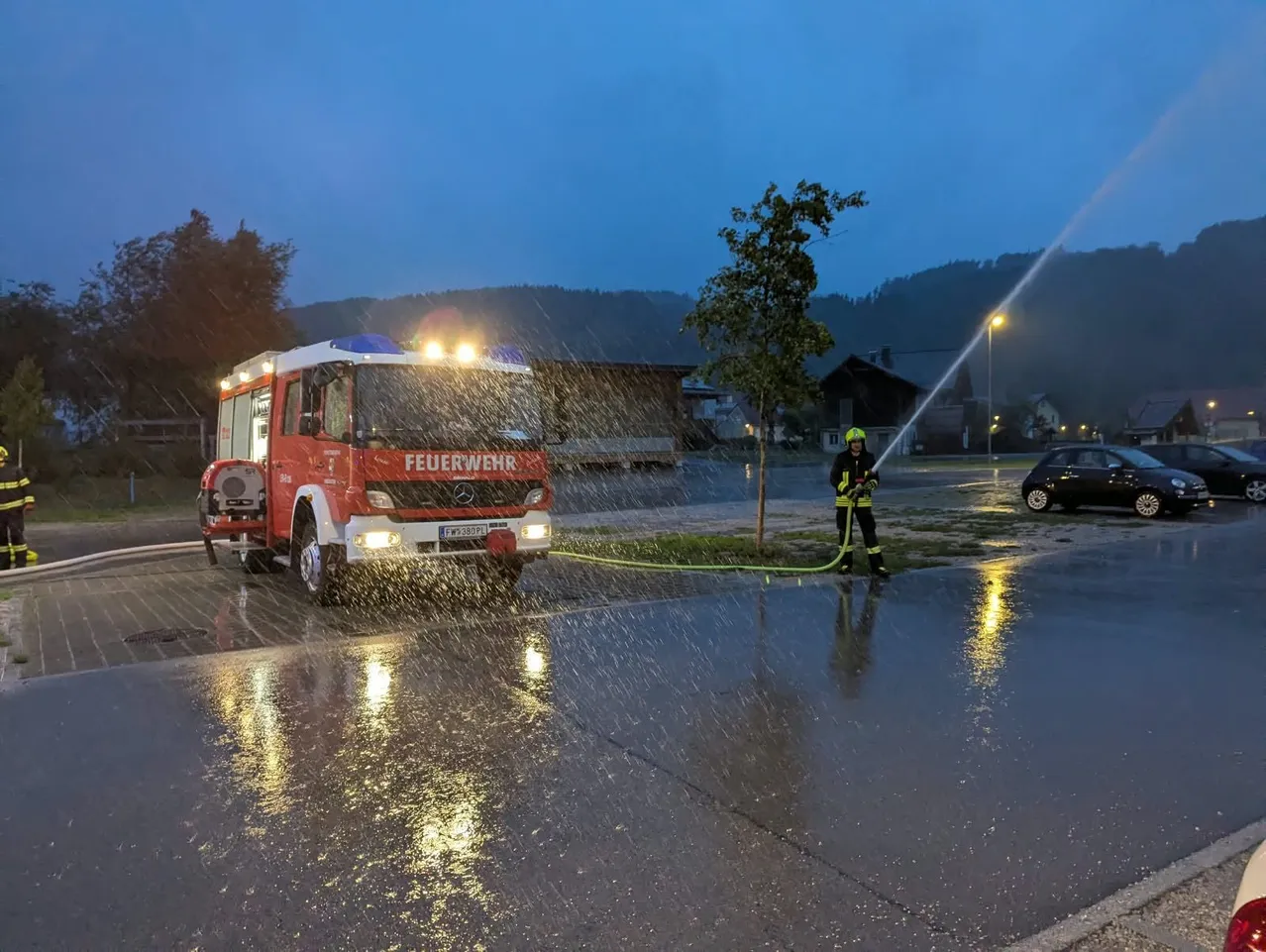 Ein Feuerwehrmann in einem gelben Helm sprüht Wasser aus einem Feuerwehrwagen bei Regen in der Nacht.