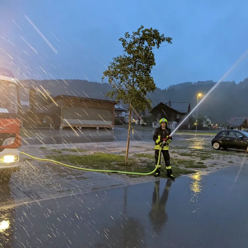 Ein Feuerwehrmann steht im Regen und sprüht Wasser aus einem Schlauch. Ein roter Feuerwehrwagen und ein kleines Auto sind in der Nähe.