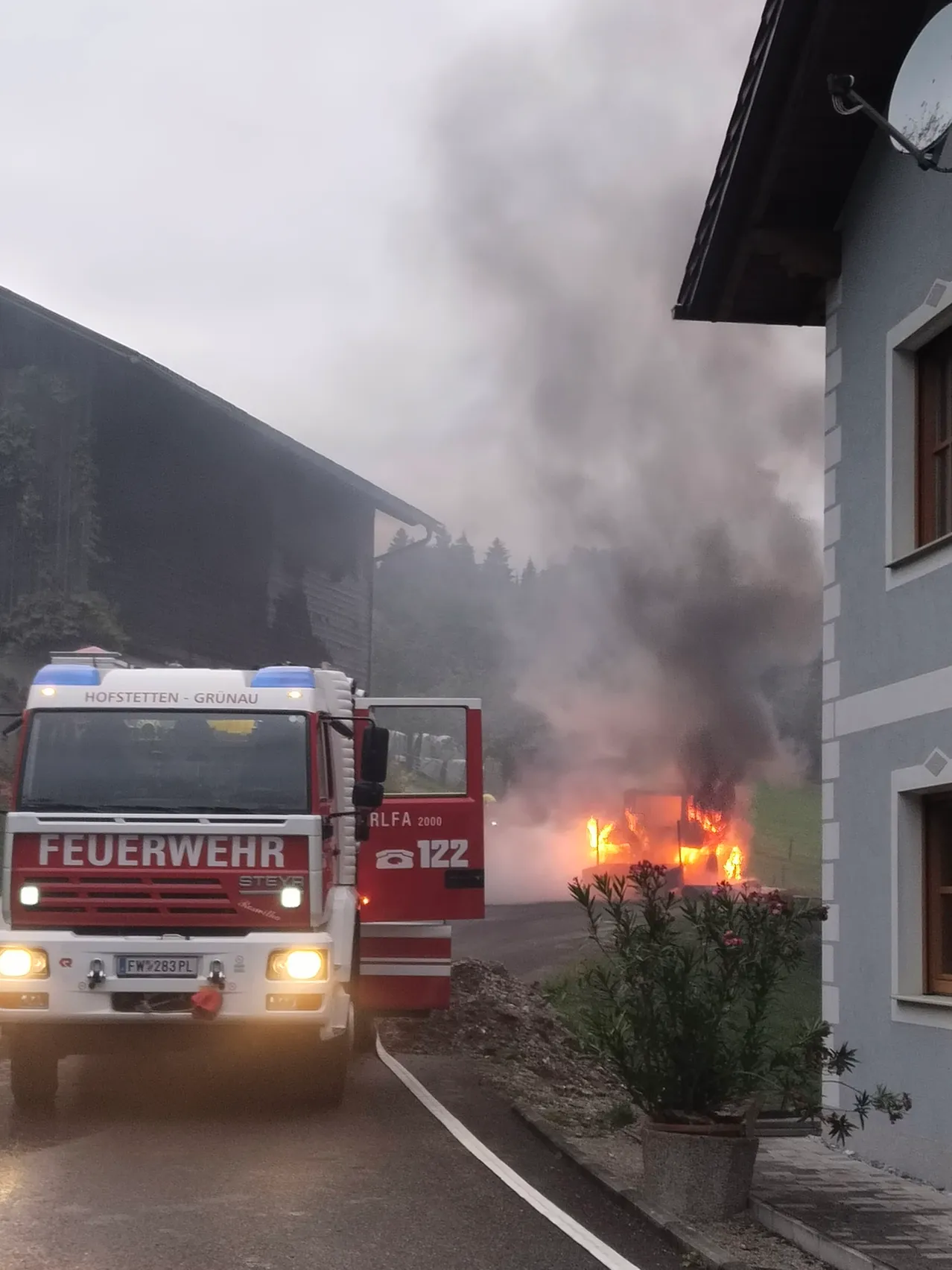 Ein Feuerwehrauto mit der Aufschrift 'Feuerwehr' steht am Straßenrand, aus einem brennenden Gebäude steigt Rauch auf.