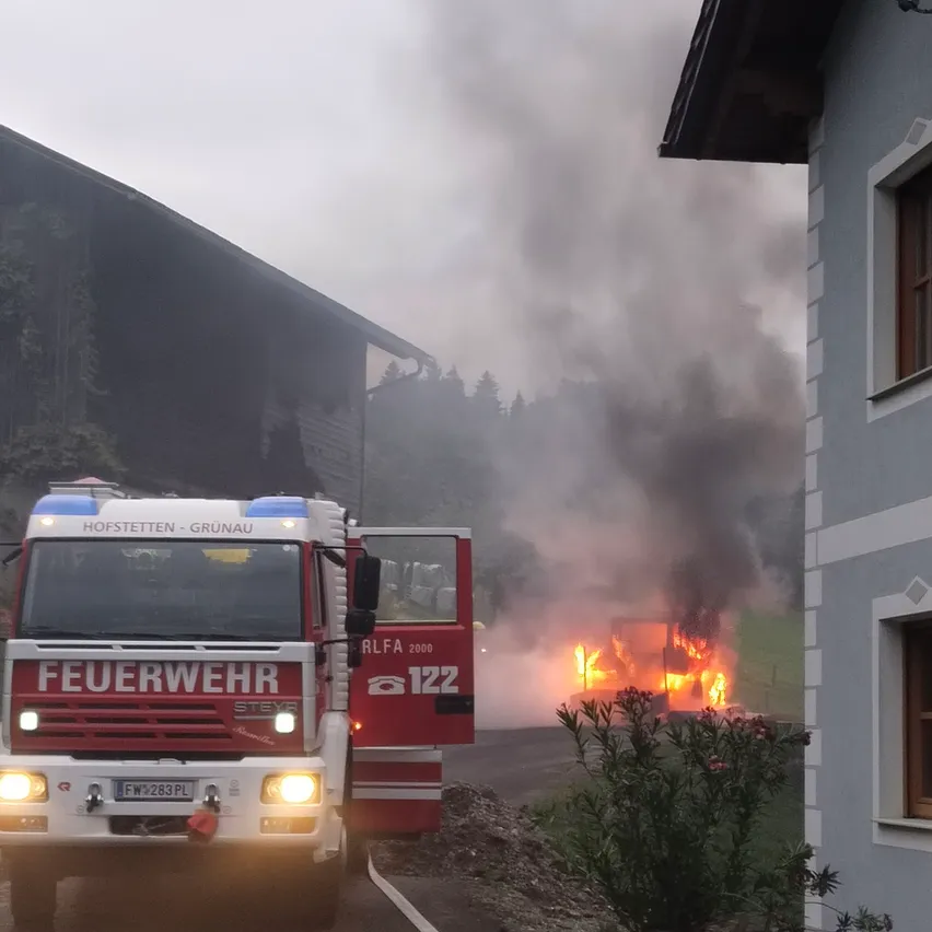 Ein Feuerwehrauto mit der Aufschrift 'Feuerwehr' steht am Straßenrand, aus einem brennenden Gebäude steigt Rauch auf.