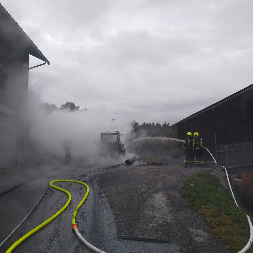 Feuerwehrleute löschen mit einem Schlauch ein Feuer vor einem Gebäude. Rauch steigt von der Szene auf.