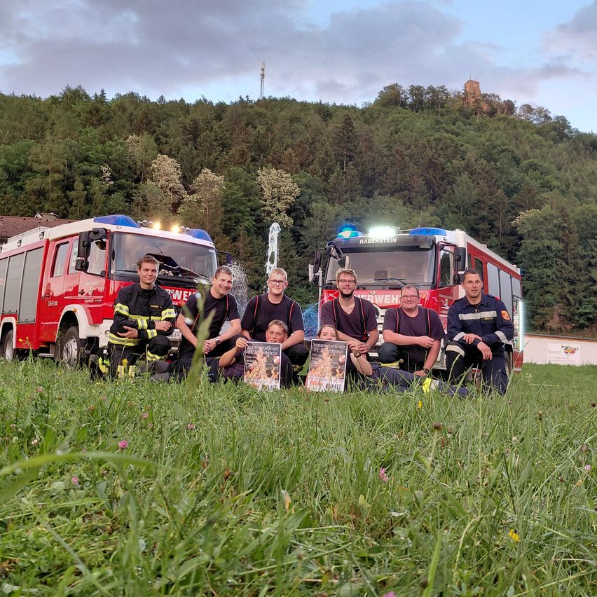 Feuerwehrleute posieren für ein Bild mit Feuerwehrautos im Hintergrund unter einem bewölkten Himmel. Sie tragen alle Uniformen und einige halten Broschüren.