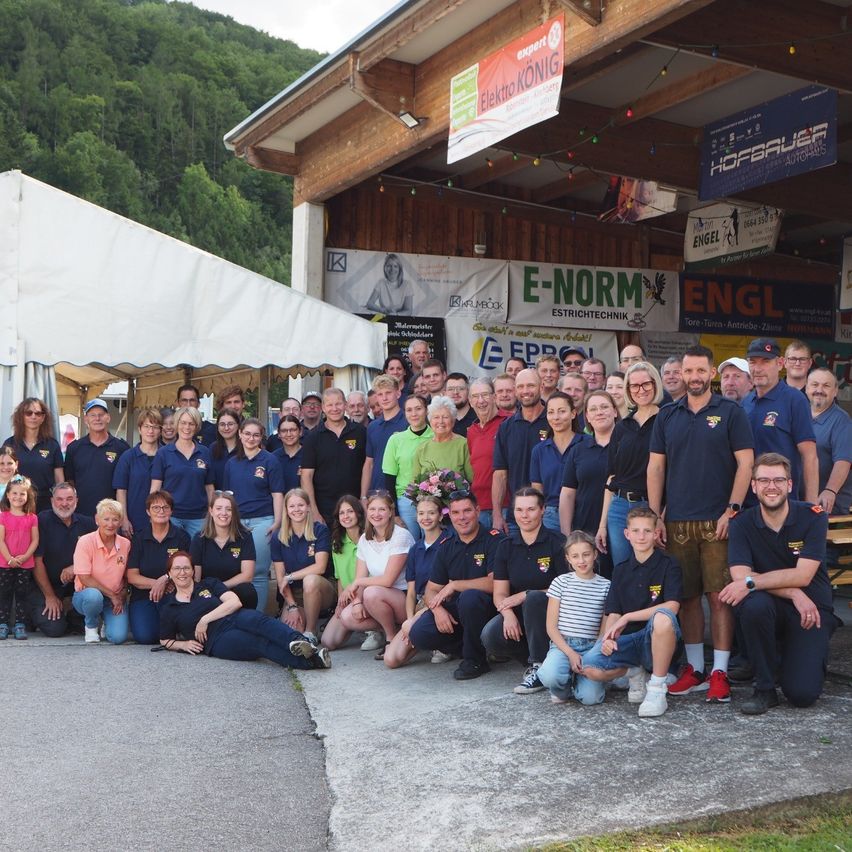 Eine Gruppe von Menschen in blauen Uniformen, einige mit Brillen, Hüten und Turnschuhen, posiert für ein Foto vor einem Gebäude mit Bannern und einem Zelt.