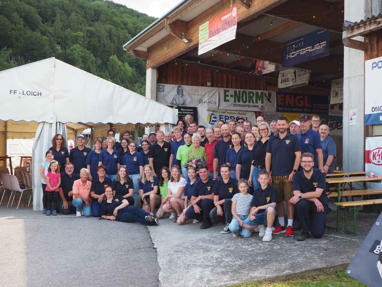 Eine Gruppe von Menschen in blauen Uniformen, einige mit Brillen, Hüten und Turnschuhen, posiert für ein Foto vor einem Gebäude mit Bannern und einem Zelt.