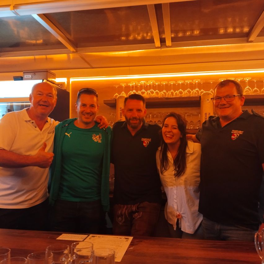 Fünf Personen posieren in einer Bar mit einer Theke und Gläsern vor ihnen. Ein Mann in Grün trägt eine Jacke mit Logo.
