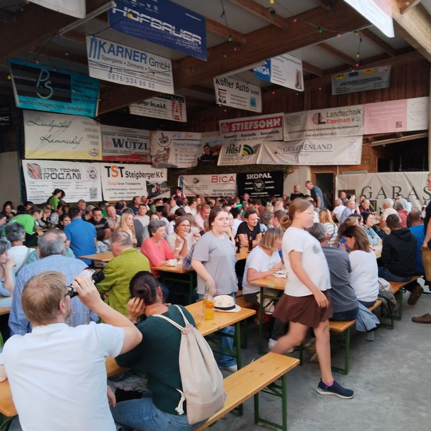 Eine Menschenmenge sitzt an Tischen in einer großen überdachten Halle. Werbung hängt von der Decke, und die Leute essen und trinken. Der Boden ist aus Beton.