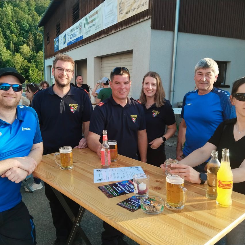 Eine Gruppe von Menschen in Uniformen steht hinter einem Holztisch im Freien und posiert lächelnd für ein Foto mit Biergläsern und Flaschen auf dem Tisch.
