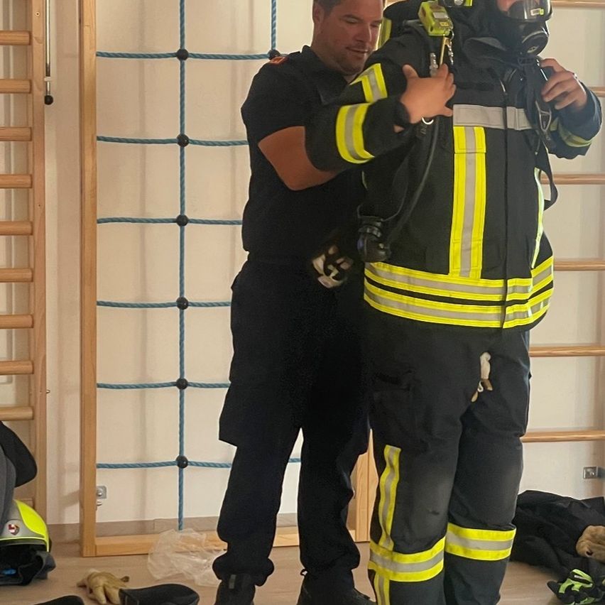 Ein Feuerwehrmann justiert die Ausrüstung eines anderen Feuerwehrmanns in einem Schulungsraum, mit einem blauen Seilleiter im Hintergrund.
