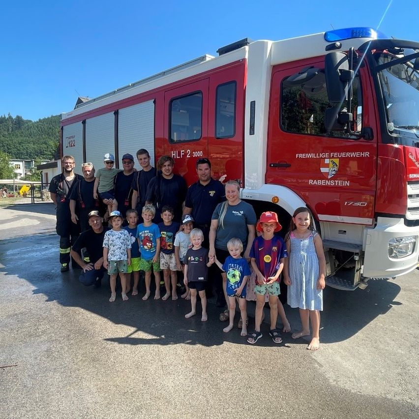 Eine Gruppe von Feuerwehrleuten und Kindern posiert vor einem roten Feuerwehrauto mit der Aufschrift 'Freiwillige Feuerwehr Rabenstein' an der Seite.