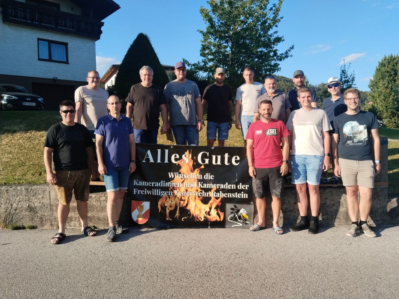 Eine Gruppe von Männern steht vor einem Banner mit der Aufschrift 'Alles Gute' und einem Feuerdesign, posiert für ein Foto. Sie sind lässig gekleidet, einige tragen Hüte und Sonnenbrillen. Hinter ihnen sind ein Haus und Bäume zu sehen.