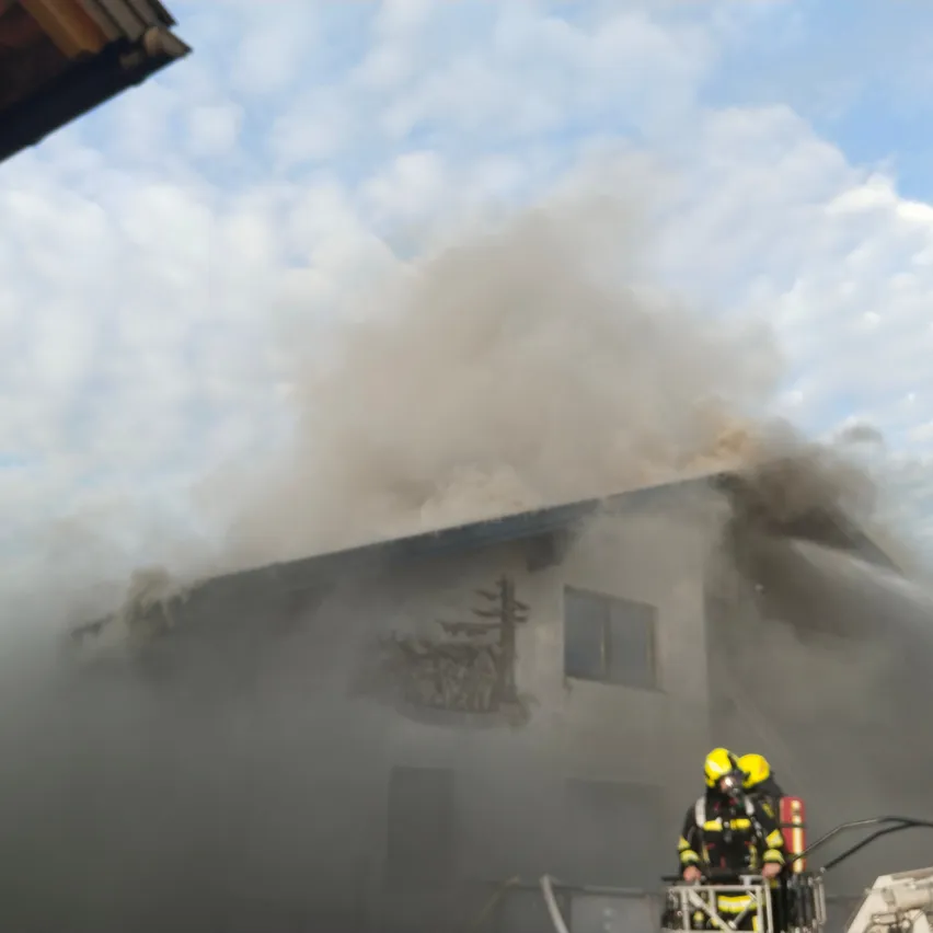 Feuerwehrleute in Schutzkleidung sprühen Wasser auf ein brennendes Haus. Rauch steigt aus dem Dach und den Fenstern auf.
