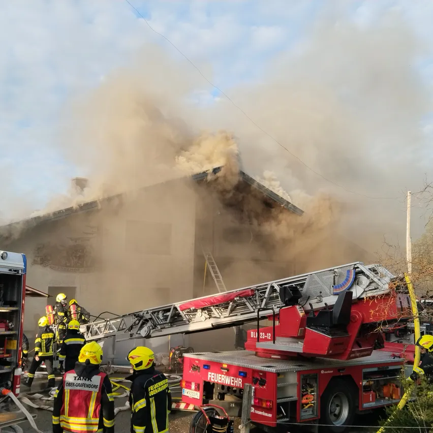 Feuerwehrleute bekämpfen einen Brand in einem Wohngebäude, aus dem Rauch aufsteigt. Ein roter Feuerwehrwagen mit ausgefahrener Leiter steht in der Nähe.