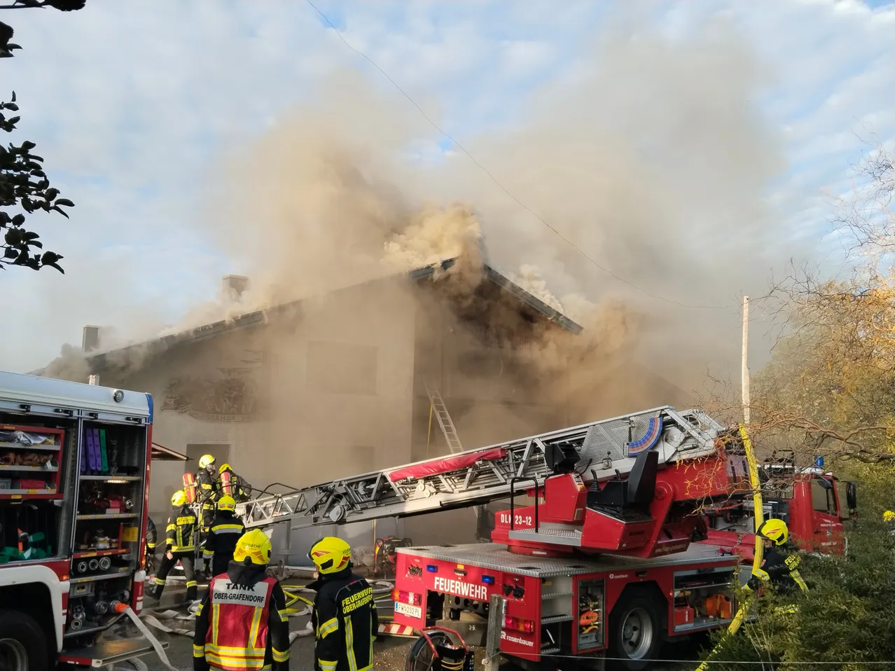 Feuerwehrleute bekämpfen einen Brand in einem Wohngebäude, aus dem Rauch aufsteigt. Ein roter Feuerwehrwagen mit ausgefahrener Leiter steht in der Nähe.