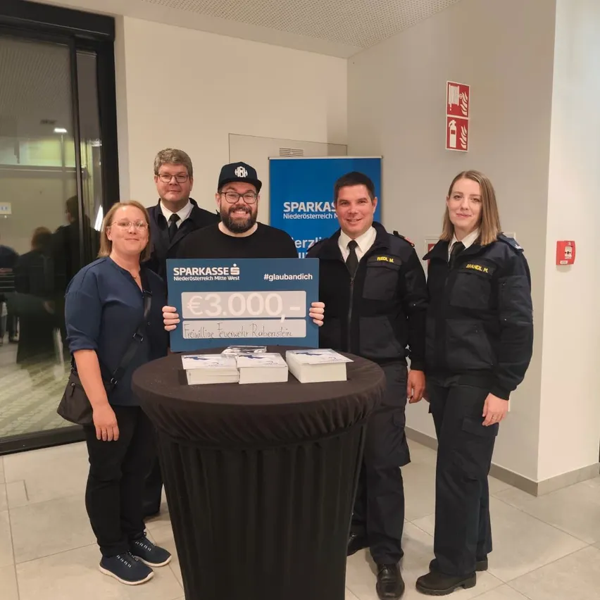 Eine Gruppe von Menschen steht vor einem Tisch mit einem blauen Banner und einem Schild mit der Aufschrift '3.000 €'.