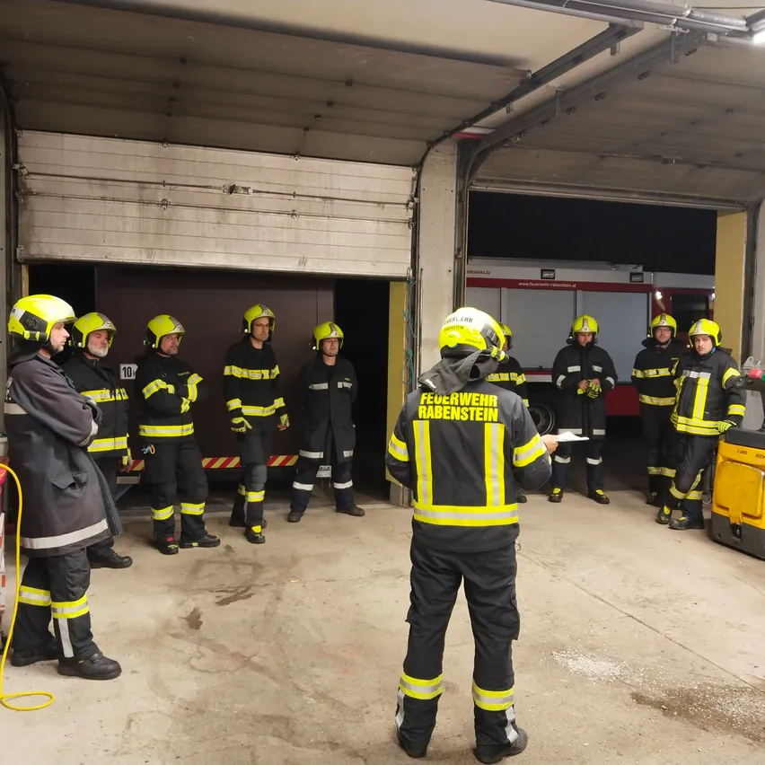 Eine Gruppe von Feuerwehrleuten in einer Garage, einer von ihnen hält eine Rede, während die anderen zuhören.