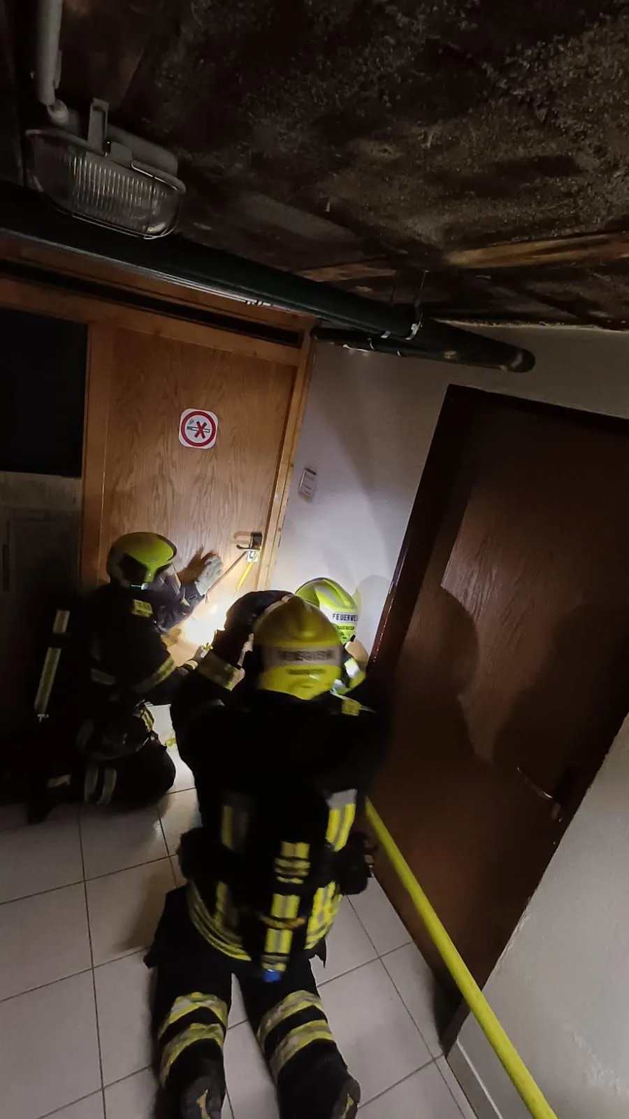 Drei Feuerwehrleute in Schutzkleidung arbeiten an einer Tür in einem Gebäude. Einer kniet und scheint etwas zu reparieren, während die anderen beiden stehen und ihn ansehen.