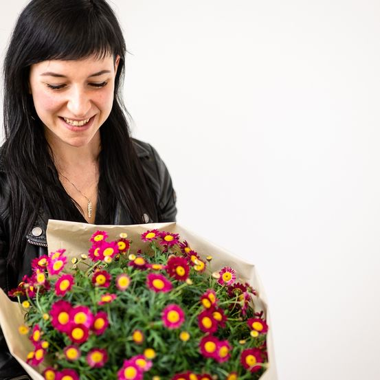 Eine Frau mit langen schwarzen Haaren und einer schwarzen Lederjacke hält einen Strauß pinker und gelber Blumen.